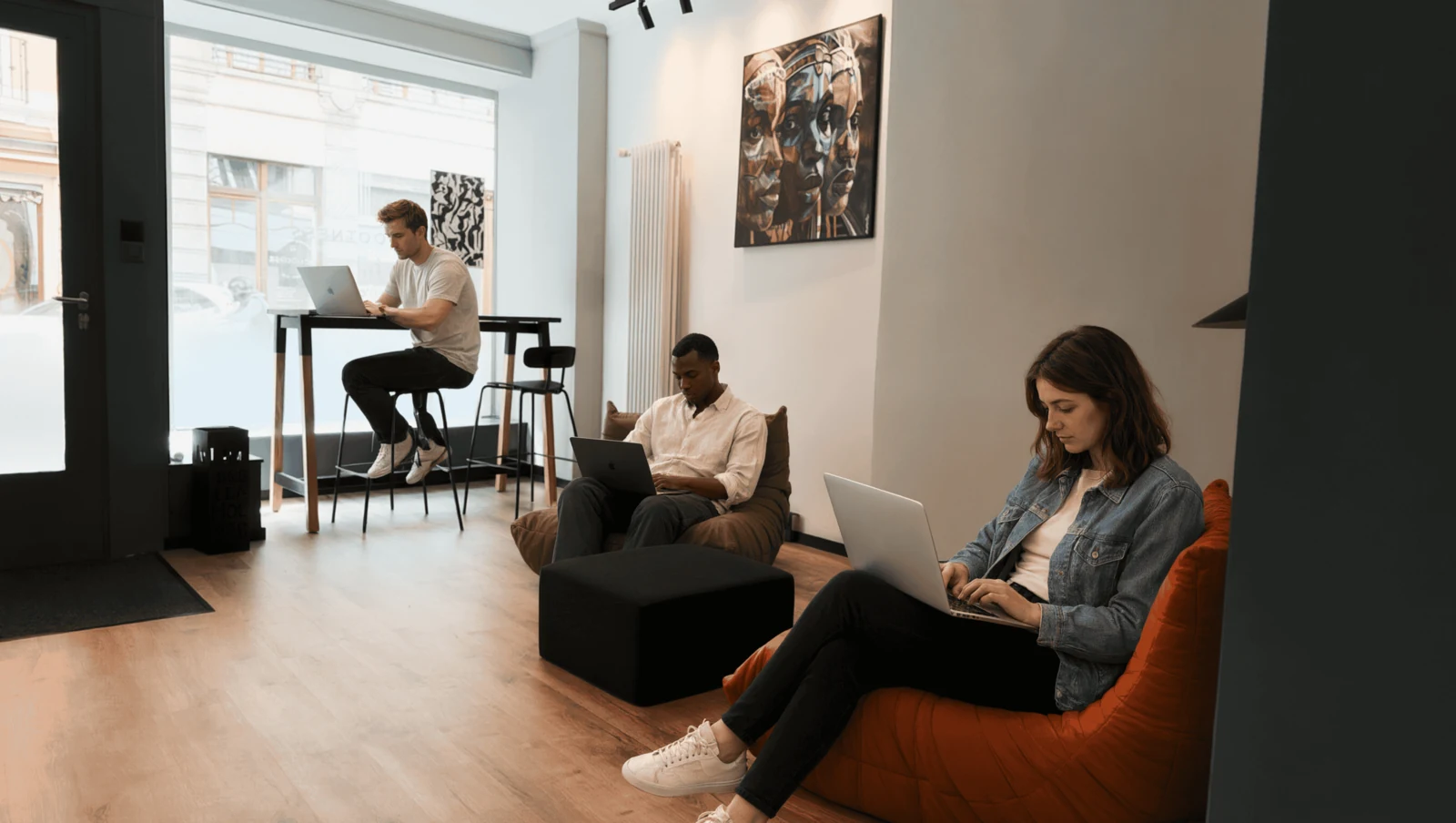 Espace détente du studio : trois personnes travaillent sur leur ordinateur portable autour d’un pouf noir et d’un fauteuil orange